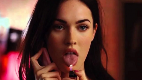 Jennifer, played by Megan Fox, holds a phone up to one ear and lit lighter in her other hand with the flame burning he end of her tongue.